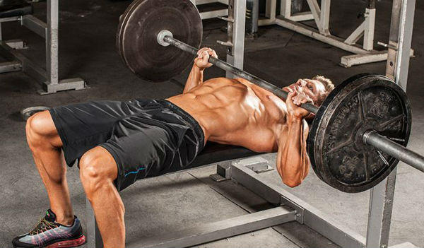 A man in black shorts lying on a bench with a barbell on his chest while performing on a bench press of 225 pounds. 