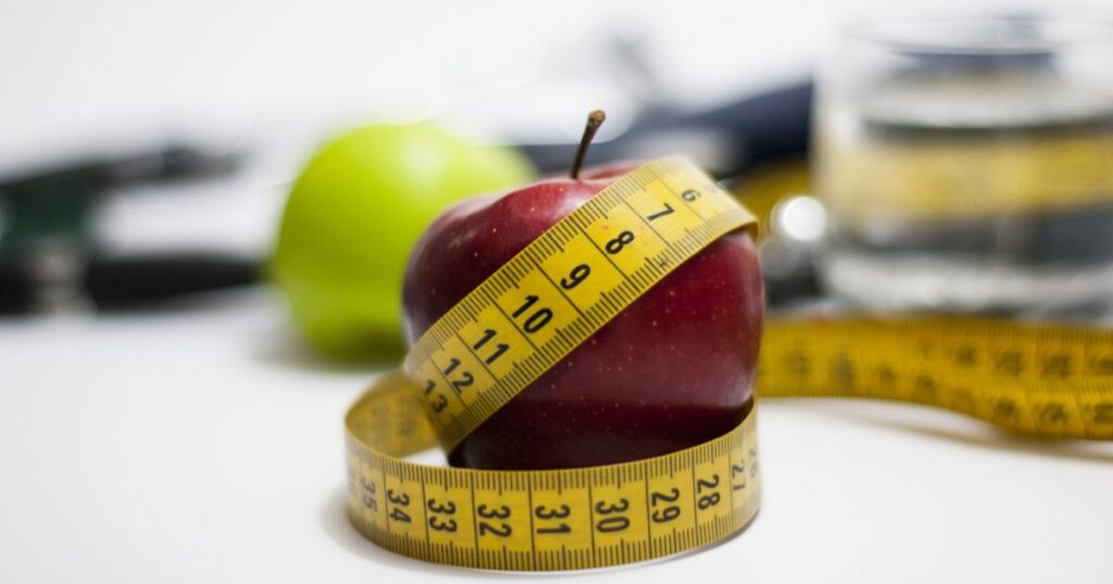 Green granny smith is in the background with a red delicious apple in the foreground that has a measuring tape around it.