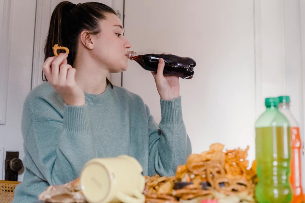 A woman stress eating pretzels and junk food while debating if an emotional eating coach is right for her.