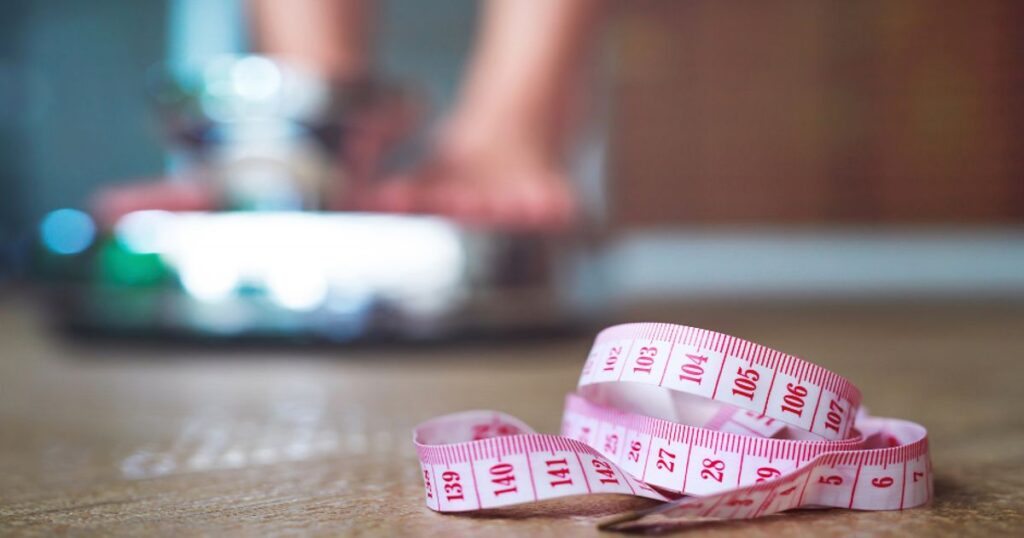 A person is measuring themselves on a scale with a measuring tape in the foreground to symbolize overcoming a sticking point. 