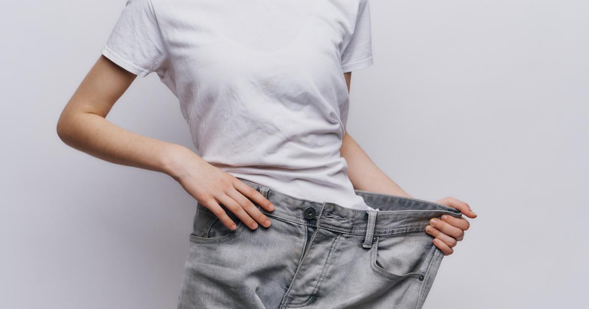 A woman is holding a large pair of jeans next to her waist, and they no longer fit because she followed the 3 steps to lose weight and kept the weight off for good.