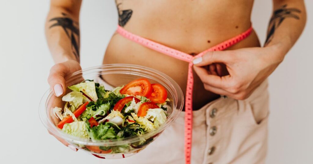 A woman is holding a measuring tape around her stomach while holding a bowl full of lettuce and tomatoes. 