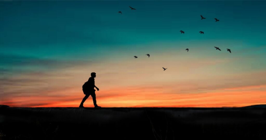 A man's silhouette is walking as the sun sets, and there are shades of red and blue in the sky. 