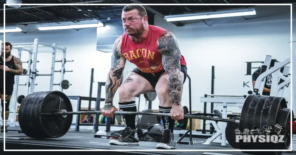 A man in a red shirt is using muscles in his posterior chain to deadlift 545 pounds using a hook grip. 