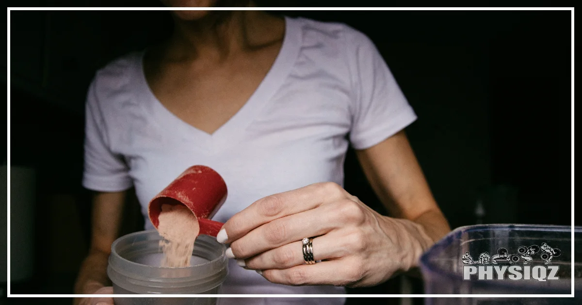 A woman wearing a white v-neck t-shirt with a ring finger, pours a chocolate-flavored protein powder into a clear tumbler in a dimly-lit room.