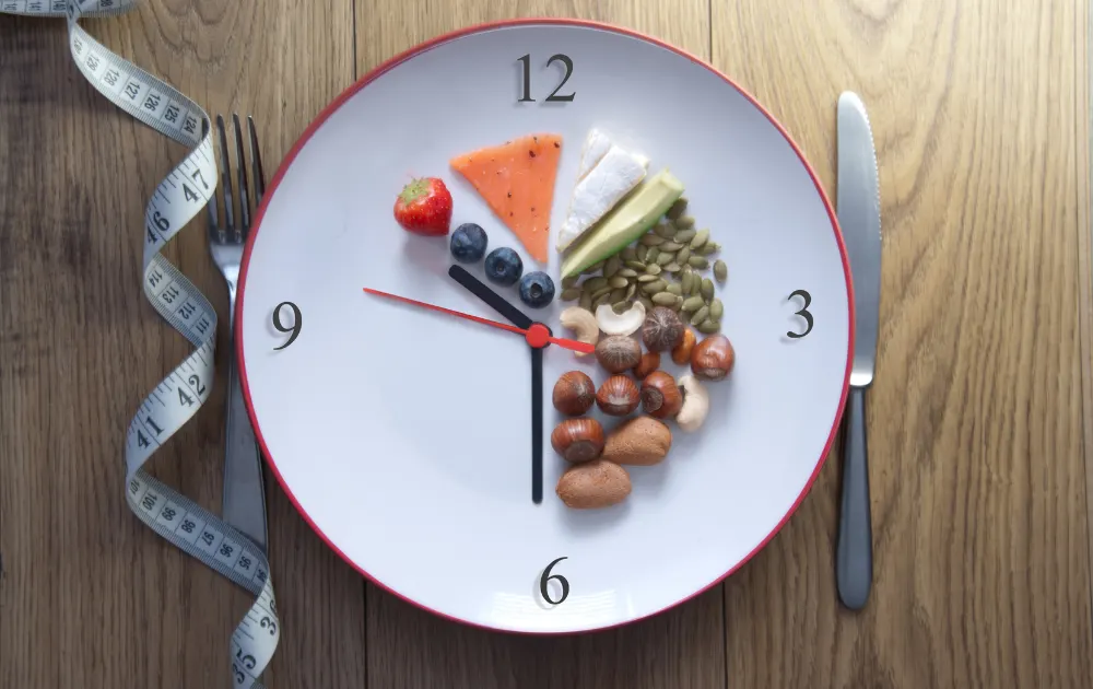 A plate with strawberries, blueberries, nuts, pumpkin seeds, cream cheese, and avocado, accompanied by a tape measure, knife, and fork, features a 24-hour clock in the center to symbolize various intermittent fasting methods like alternate day fasting and one meal a day (OMAD).