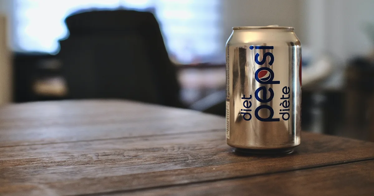 A shiny bronze can of diet Pepsi placed on top of a wooden table in a room with blurred chairs in the background.