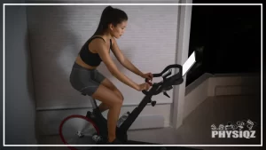 A woman wearing a black tank top, grey shorts and black shoes exercising using a Peloton bicycle while following the best peloton class for weight loss training video displayed on the gadget included in the bike, in a dimly-lit room.
