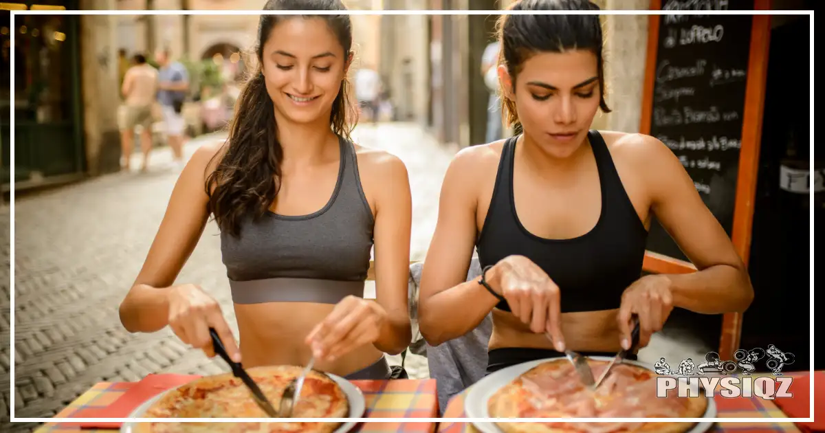 Two women wearing fitness clothes after a workout are eating and cutting into pizza on a cheat day while sitting in the middle of a city street, wondering, "Can you eat whatever you want if you workout?" or does it matter what you eat?