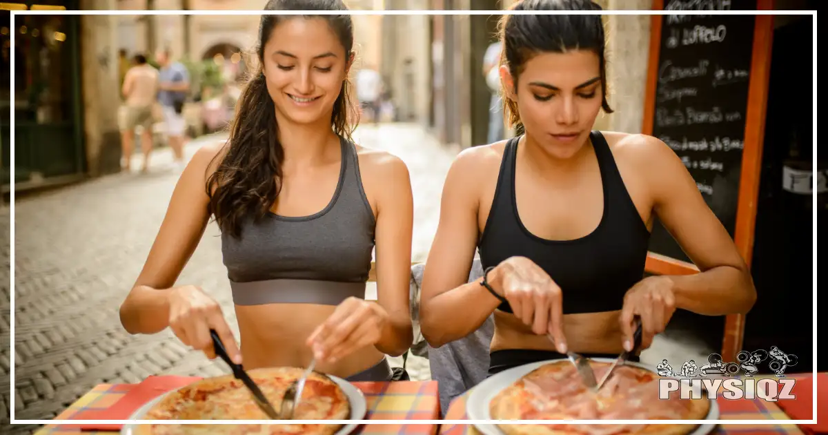 Two women in fitness attire enjoy pizza on a cheat day in the middle of a city street, pondering the impact of their food choices on their workout results.