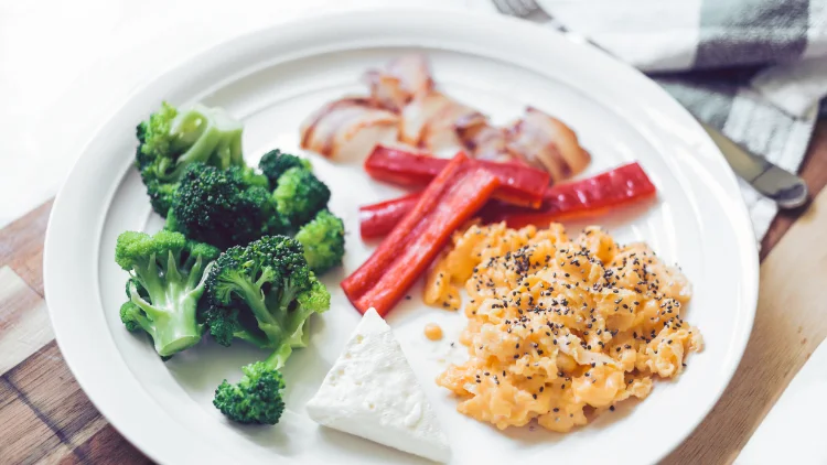 A white dish full of broccoli, scrambled eggs, bacon and bell peppers on a wooden tray. 