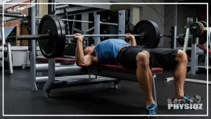 A man wearing a blue shirt and black shorts is laying on a bench pressing up a barbell with two plates on it which made him ask himself "How many people can bench 225?"