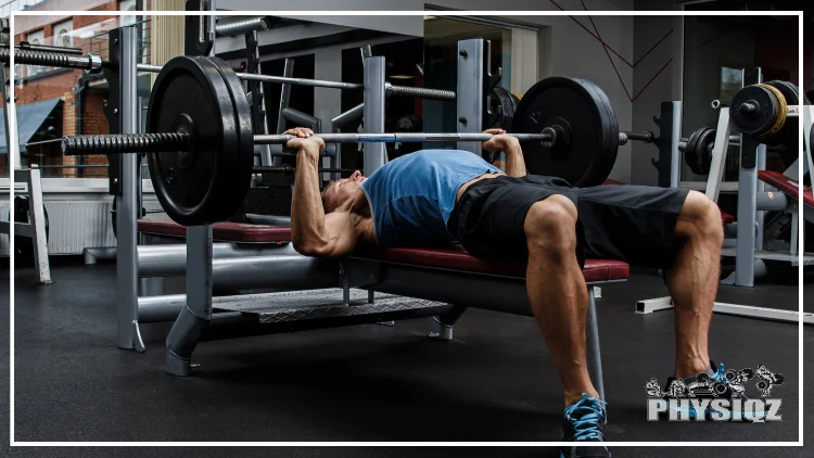 A man in a blue shirt and black shorts is lying on a bench, pressing a barbell with two plates on each end.