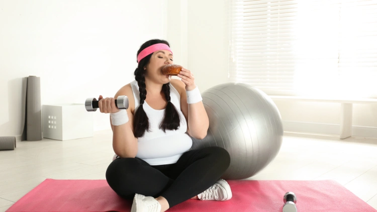 An overweight woman with a pink headband, wearing a white tank top, black leggings, and white shoes sitting on a pink yoga mat, while eating a bun and holding a silver dumbbell on the other hand, and the other pair of the dumbbell is placed on the yoga mat, and a gray exercise ball can be seen behind her.