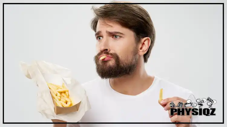 A man is holding a cardboard box full of carb-rich french fries in his right hand, is eating a fry with his left hand, and has a face of uncertainty. 