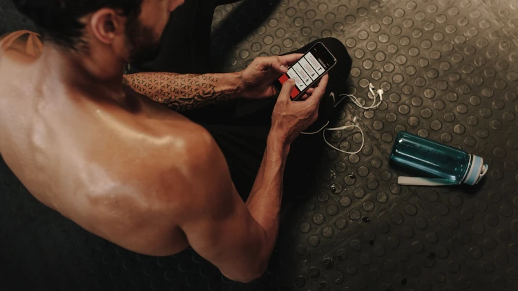 A shirtless man with beard and a tattoo on his left arm is monitoring his workout progress using a fitness app on his phone during his break in a gym, and a clear blue water bottle can be seen lying on the floor.