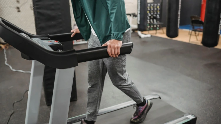 A man wearing a green jacket with a white t-shirt under, grey sweat pants, and shoes with black socks is walking on a treadmill in a gym with punching bags in the background.
