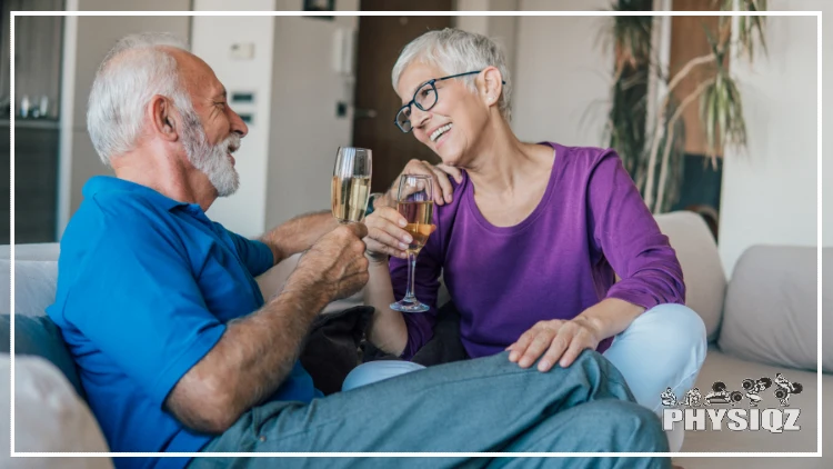 A couple with grey hair is sitting on a couch while each is holding a glass of wine, smiling, and relieving stress from the day. 