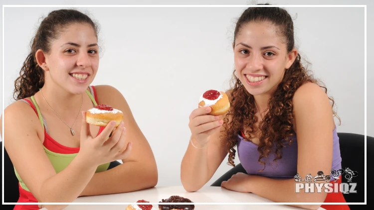 Two girls in tank tops are sitting at a table holding cream filled donuts that are glazed and covered in powdered sugar. 