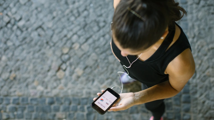 A tan woman with earphones on, wearing a black tank top and pants is checking a fitness app on her phone, monitoring her workout progress.