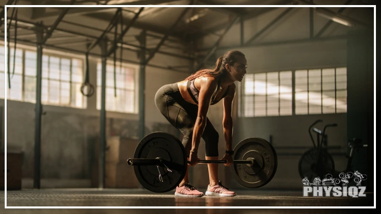 A woman in warehouse is wearing pink shoes, black shorts and a tank top as she gets into the bottom of a deadlift with a double overhand grip 
