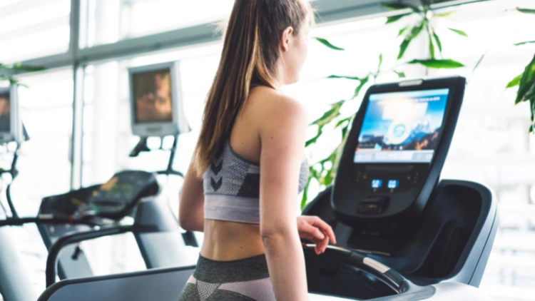 A woman wearing a matching grey tank top and pants using a Peloton treadmill while watching a fitness class in a gym with other treadmills in the background.