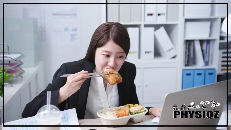 An Asian woman in an office and at her desk as she eats fried food with chop sticks and has a milkshake next to her. 