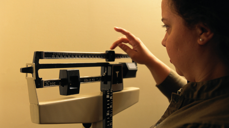 A woman with strawberry blonde hair is looking at a physician's scale with a face of concern and curiosity as she finalizes her weight, which shows she's 192 pounds or heavier than expected. 
