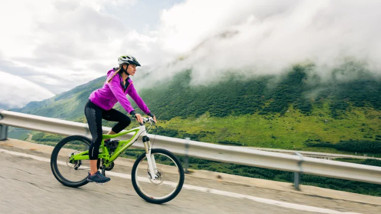 A young cyclist woman wearing a purple windbreaker jacket, black leggings and shoes, with black helmet on, is riding a bike with a view of the mountainside in the background.