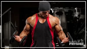 A vascular and muscular guy is wearing a black beanie and a black tank top with red stripes on the side is looking down at his right bicep as he performs a bicep curl which is a part of the 5 day powerbuilding split workout routine he's following.