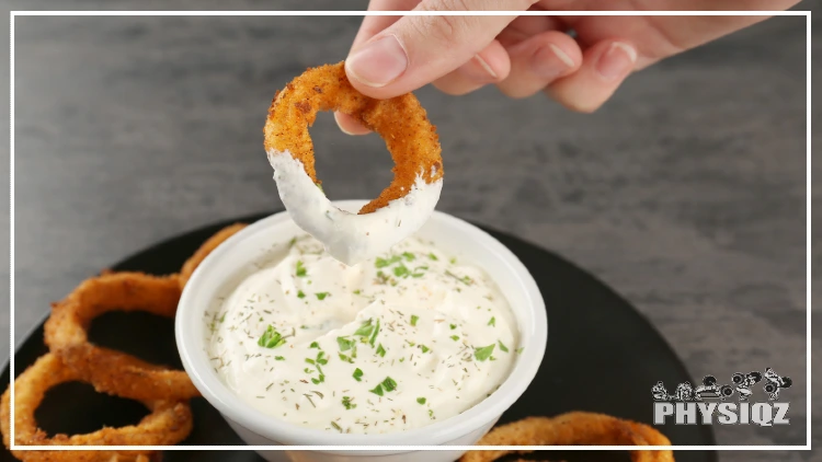 A person's pointer and thumb are grasping a crispy opinion ring that's orange-yellow with the bottom half covered in ranch as they pull it out of a ranch cup that contains green chives, which makes them question, "Are onion rings good for weight loss?".