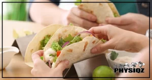 Two pairs of hands are holding tacos made with carb balance tortilla keto recipe and contain lettuce, onions, tomatoes and shrimp while some tacos are placed in a silver taco plate set beside a lime and a cup of sour cream.