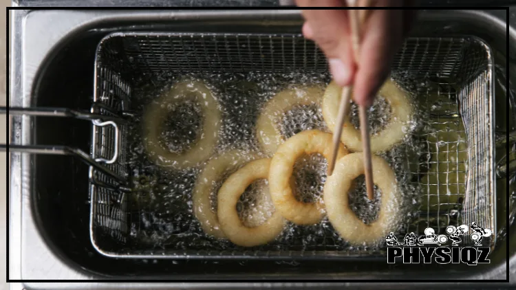 A deep fryer basket is submerged in sizzling vegetable oil and has seven light yellow onion rings being cooked while someone flips the rings from the top with chop sticks. 
