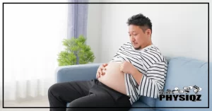 A man sitting a light blue couch in a white colored room wearing a white and black striped shirt and black jeans is rubbing his round stomach and gazing at it showing an unpleased expression on his face as he asks himself "Does sitting after eating make your belly fat?".