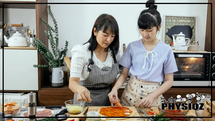 Two women, one wearing a gray apron and a white shirt and the other wearing a blue shirt and tan apron, are in a kitchen with white walls, two white teapots, a white vase with a small plant it in and a small oven while the women are in the middle of making a pizza as there is an array of pizza toppings on the counter like pepperoni, cheese, salami and black pepper.