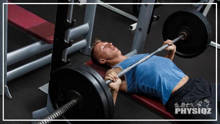 A man wearing a blue tank top and black shorts is laying on a red bench at the gym with his back arched, holding onto a barbell with two 45 pound weights on either end and is in the staring position of a bench press with the barbell touching his mid chest.