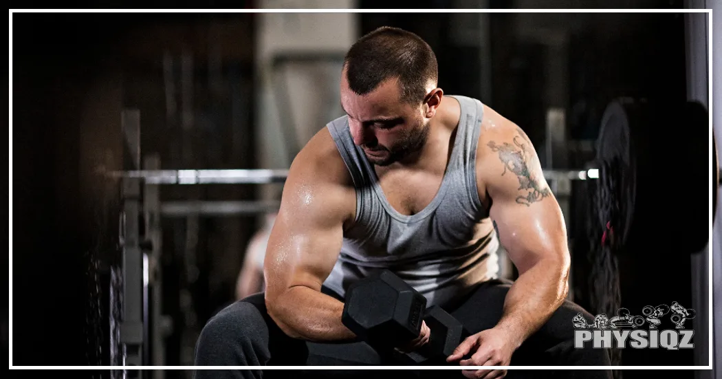 A man wearing a gray tank top and black pants is in a gym with a blurred background and sitting on a bench with both elbows on his knees while holding a dumbbell as he performs a bicep curl to target the hardest muscle to grow for himself.