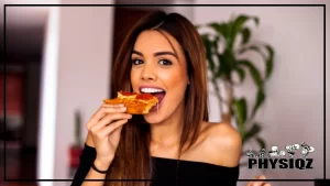 A woman wearing a black, off the shoulder shirt is standing in front of a white wall and a green house plant while biting into a slice of pepperoni pizza and wondering, "Is Mod Pizza Cauliflower Crust Keto Friendly?".