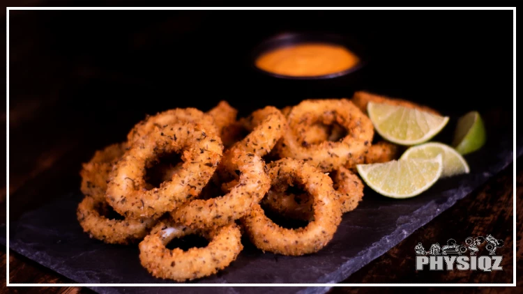 A square black plate is holding golden brown onions rings with black dots of seasoning over them and there's four bright green lime slices on the right and an orange dipping sauce in the background. 