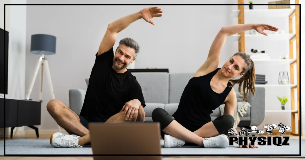 A male and a female, both wearing all black workout attire, are sitting crisscrossed and stretching their arms and torso's to the right while in a bright living room watching their laptop and performing their Peloton workout.