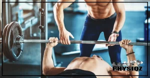 A man in black shorts is wondering where should the bar touch on bench press as he lays on a bench in the middle of a gym while pressing up on a 225 pound bench press with another guy in blue shorts spotting him from behind.
