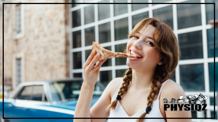A woman is outside in front of a blue vehicle, wearing her hair in 2 braids and in a white tank top while she is smiling and taking a bite of a slice of pizza.