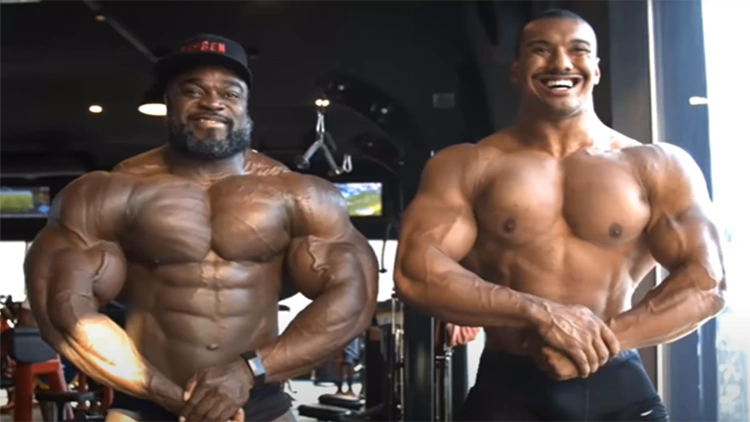 Brandon curry, a black bodybuilder is shown flexing on the left with gigantic muscle size and striations on his shoulders and quads since he's so lean while Larry Wheels, a professional powerlifter is on his right in the same pose but his muscles are much smaller and not as aesthetically pleasing since his upper body is disproportionately larger than his lower body and there's red and black gym equipment in the background. 