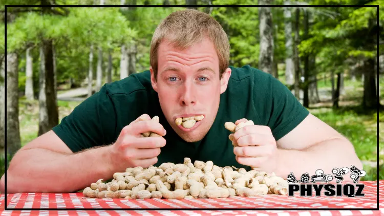 A Caucasian man wearing a green t-shirt is stuffing his mouth with boiled peanuts from the table with checkered tablecloth while thinking are boiled peanuts keto, in an outdoor setting with trees in the background.