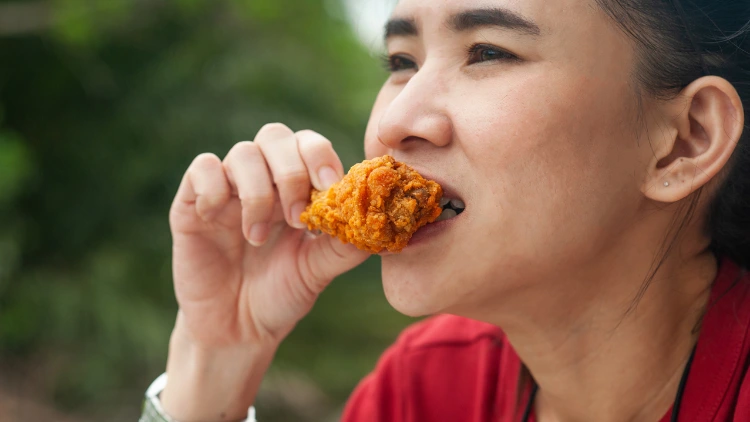 An Asian woman wearing a red t-shirt is eating a piece of fried chicken wings outdoors.