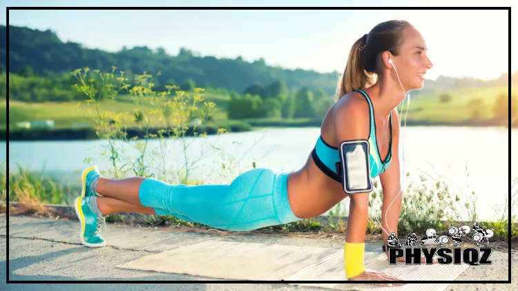 A woman in blue pants, a top, and shoes has her feet crossed, chest up, and arms underneath her as she does a plank variation next to a lake and grassy hills during an early morning sunrise because she knows the best time to exercise to lose belly fat for women.