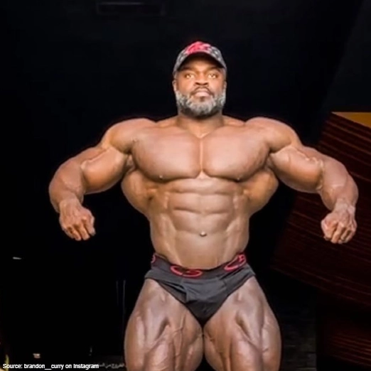 Brandon Curry, a black bodybuilder is flexing his lats while wearing a black and red snap back and his pose makes his waist appear smaller, shoulders wider, and back thicker which makes up a v-taper that's highly desired in bodybuilding. 