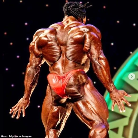 Kai Greene is wearing red swim suit bottoms as he flexes his ultra defined back that show every insertion point and there is purple, orange and pink stars in the background. 