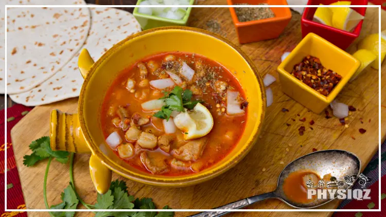 A yellow bowl of Mexican menudo that has green toppings, a lemon, and orange broth and next to is is a large silver spoon, speckled tortilla, and other condiments such as onions, chili pepper flakes, limes, and cilantro.