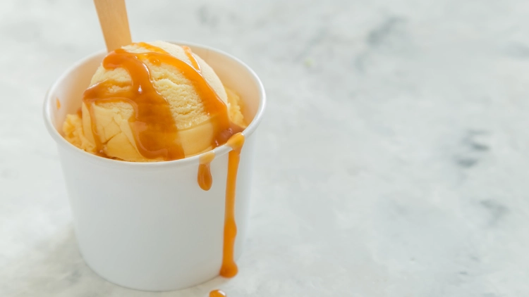 A white cup filled with caramel ice cream drizzled with caramel syrup with wooden spoon, displayed on top of a white surface.